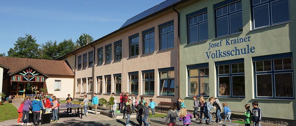 Kinder spielen vor der Josef Krainer Volksschule, einer Schule mit Solarpanelen auf dem Dach, an einem sonnigen Tag.