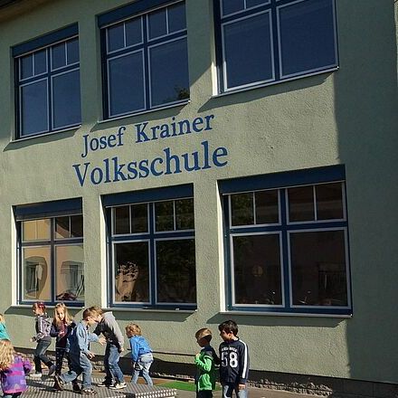 Ein Schulgebäude namens Josef Krainer Volksschule mit Solarpanels auf dem Dach, vor dem Kinder spielen. Das Gebäude hat mehrere Fenster, eine Bank und einen Tischtennistisch. Im Hintergrund sind Bäume zu sehen.