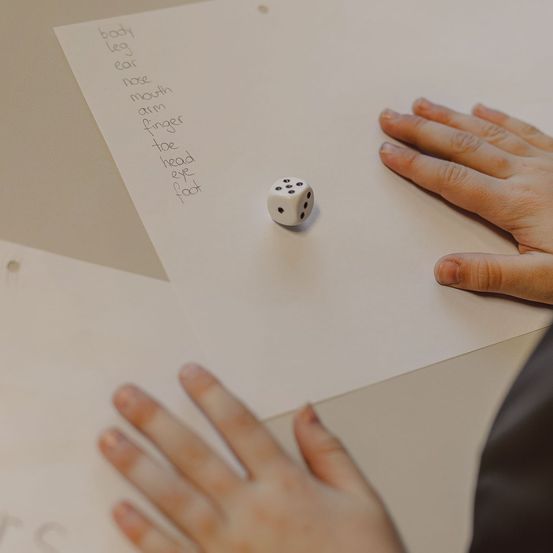 Die Hände einer Person liegen auf einem Schreibtisch und halten ein Papier mit aufgelisteten Körperteilen und einem kleinen Würfel.