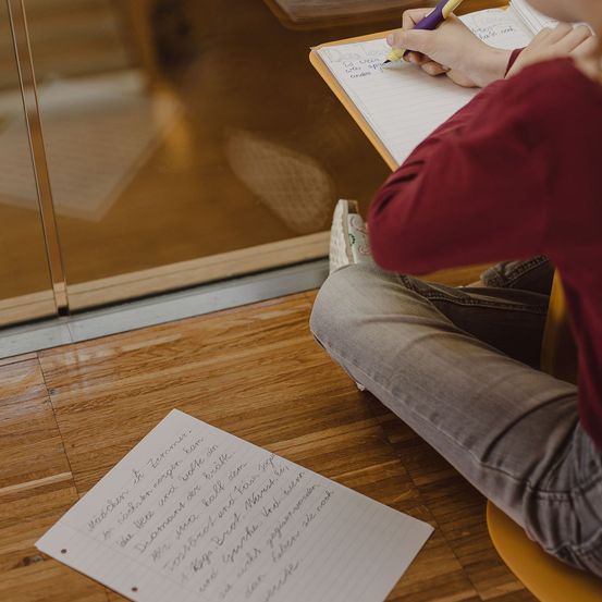 Ein Schüler sitzt auf einem Stuhl und schreibt mit einem lila Stift in ein Notizbuch. Ein Blatt Papier mit Schrift steht auf dem Boden.
