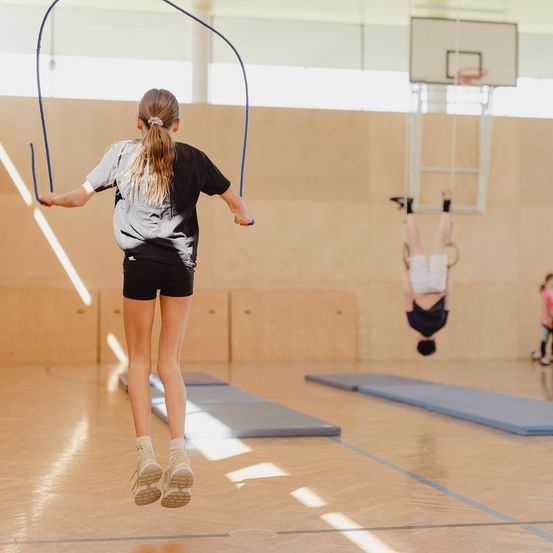 Ein Mädchen springt Seil in einer Turnhalle, während eine andere Person kopfüber an einem Reifen hängt. An der Wand ist ein Basketballkorb montiert.