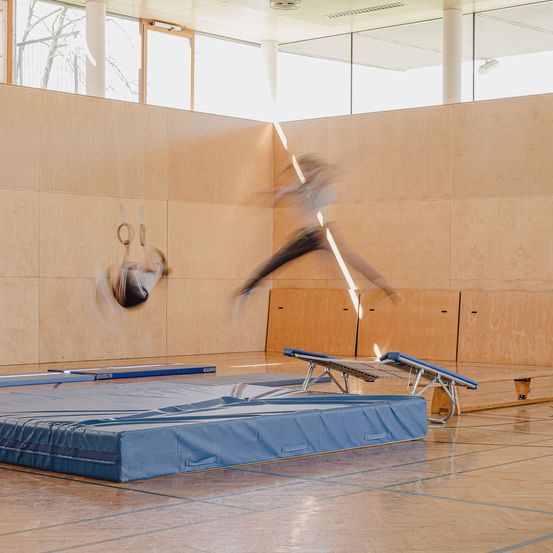 Zwei Turner in einem Fitnessstudio üben Sprünge. Einer ist in der Luft, und der andere hält sich an Ringen fest. Blaue Matten und Holzböden.