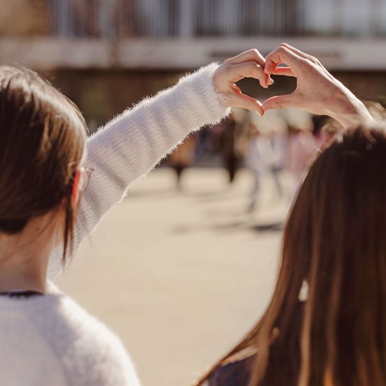 Zwei Frauen bilden mit ihren Haenden ein Herz, waehrend sie in einem offenen Bereich stehen, mit einem verschwommenen Hintergrund eines Gebaeudes.