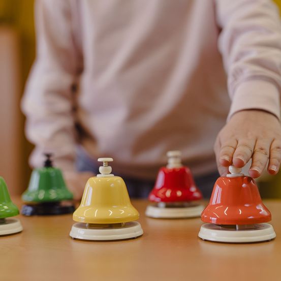 Ein Kind spielt mit einem Set farbiger Glocken auf einem Holztisch. Die Glocken sind grün, gelb, rot und orange.