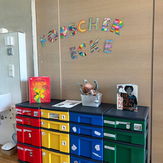 Ein Stauraum mit farbenfrohen Schubladen und Büchern, darunter ein rotes mit dem Titel Labor, neben einer Wand mit dem Wort Forscherecke in Regenbogenfarben geschrieben.
