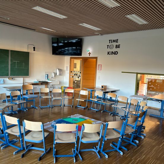 Ein Klassenzimmer mit einem Kreis aus blauen und beigefarbenen Stühlen. Ein farbenfroher Teppich liegt auf dem Boden, und eine Tafel ist an der Wand. Ein Monitor ist über einer Tür montiert, und ein motivierendes Zitat ist an der Wand angebracht.