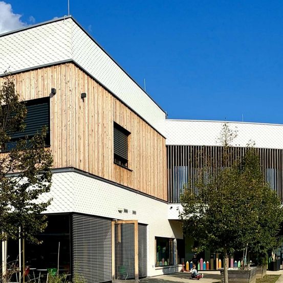 Das Äußere eines modernen Schulgebäudes mit einer Mischung aus weißen und braunen Holzpaneelen, Glasfenstern und einem großen Eingang mit einer farbenfrohen Anzeige.