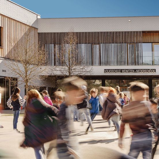 Schüler eilen an einem sonnigen Tag in das Schulgebäude der Volksschule Weinitzen-Niederschöckl. Die moderne Architektur zeigt Holz- und Glaselemente.