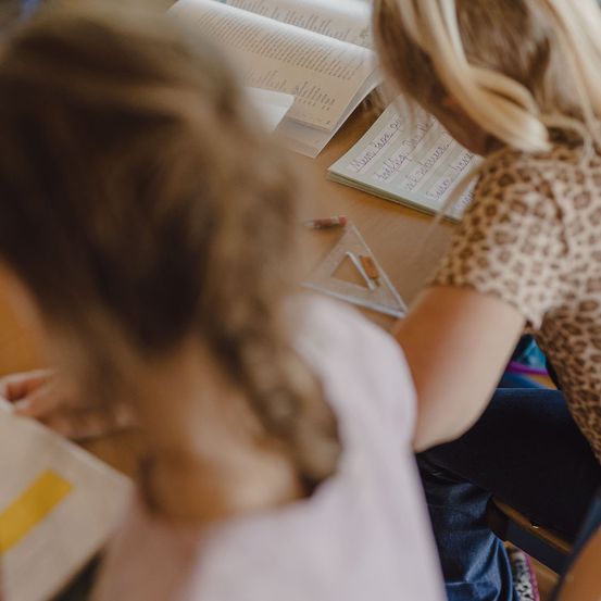 Zwei Kinder sitzen an einem Schreibtisch und beschäftigen sich mit einer Schulaktivität. Ein Kind hat ein Buch offen, und das andere schreibt auf ein Blatt Papier.