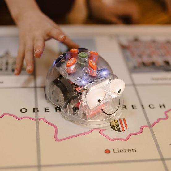 Eine Kinderhand steuert einen kleinen Roboter auf einer Karte einer Region. Der Roboter hat Knöpfe und LED-Leuchten.