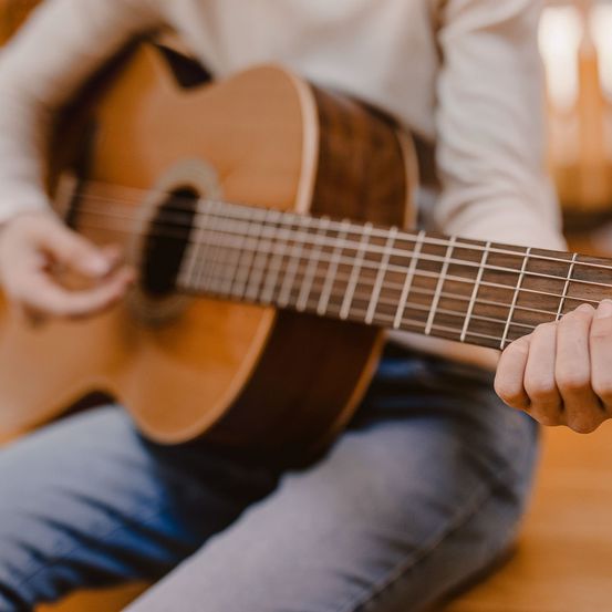 Eine Person sitzt und spielt Gitarre. Die Gitarre hat einen braunen Korpus mit weißem Plektrum. Die Hände der Person liegen auf dem Hals und Griffbrett der Gitarre.