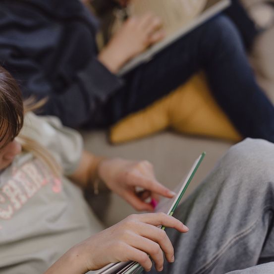 Ein junges Mädchen hält ein Buch in der Hand und sitzt auf einem Sofa, mit einer Frau hinter ihr, die einen Laptop benutzt.