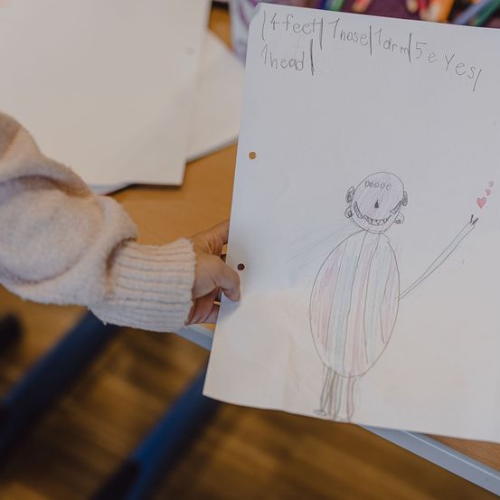 Ein Kind hält ein Papier mit einer Zeichnung einer Strichfigur hoch, über der die Körpermaße geschrieben sind.