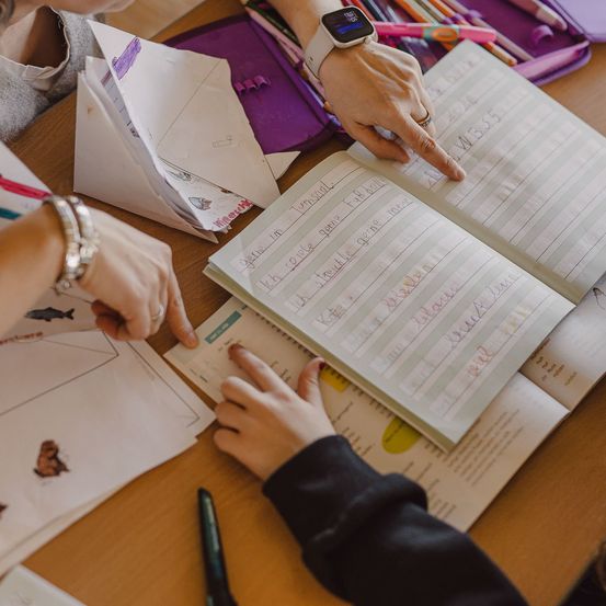 Ein Kind sitzt an einem Schreibtisch mit einem offenen Buch, und ein Lehrer zeigt auf den Text. Das Kind hat einen Stift, und es gibt Bleistifte und Marker in einem Stiftetui. Das Kind trägt eine Armbanduhr und einen Ring.
