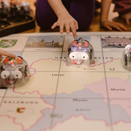Eine Kinderhand führt einen kleinen Roboter auf einer Karte mit Regionen wie Salzburg und Murtal.