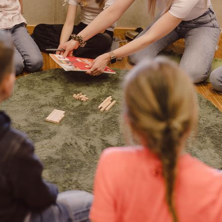 Vier junge Leute sitzen auf einem Teppich und spielen ein Spiel mit Holzteilen. Einer hält ein Buch und zeigt darauf. Ein anderer beobachtet, und zwei weitere beobachten sie.