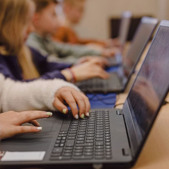 Ein Klassenzimmer mit Kindern, die Laptops benutzen. Ein Kind im Vordergrund hat die Hände auf der Tastatur.