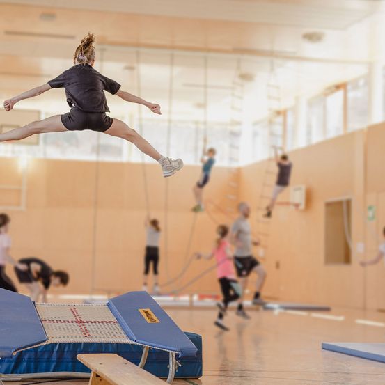 Ein Fitnessstudio mit Trainingsmatten und verschiedenen Geräten. Eine Frau springt in der Luft, während andere im Hintergrund trainieren.