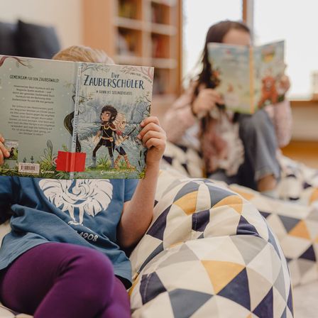 Ein Kind liest ein Buch mit dem Titel Der Zauberschüler, während ein Mädchen in der Nähe sitzt. Beide sitzen in einer gemütlichen, farbenfrohen Umgebung mit einem Bücherregal im Hintergrund.