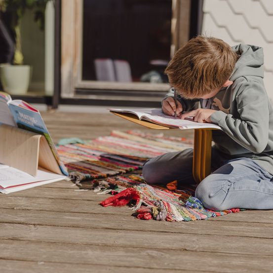 Ein junger Junge studiert auf einer Holzterrasse mit Büchern und einer Matte in der Nähe.