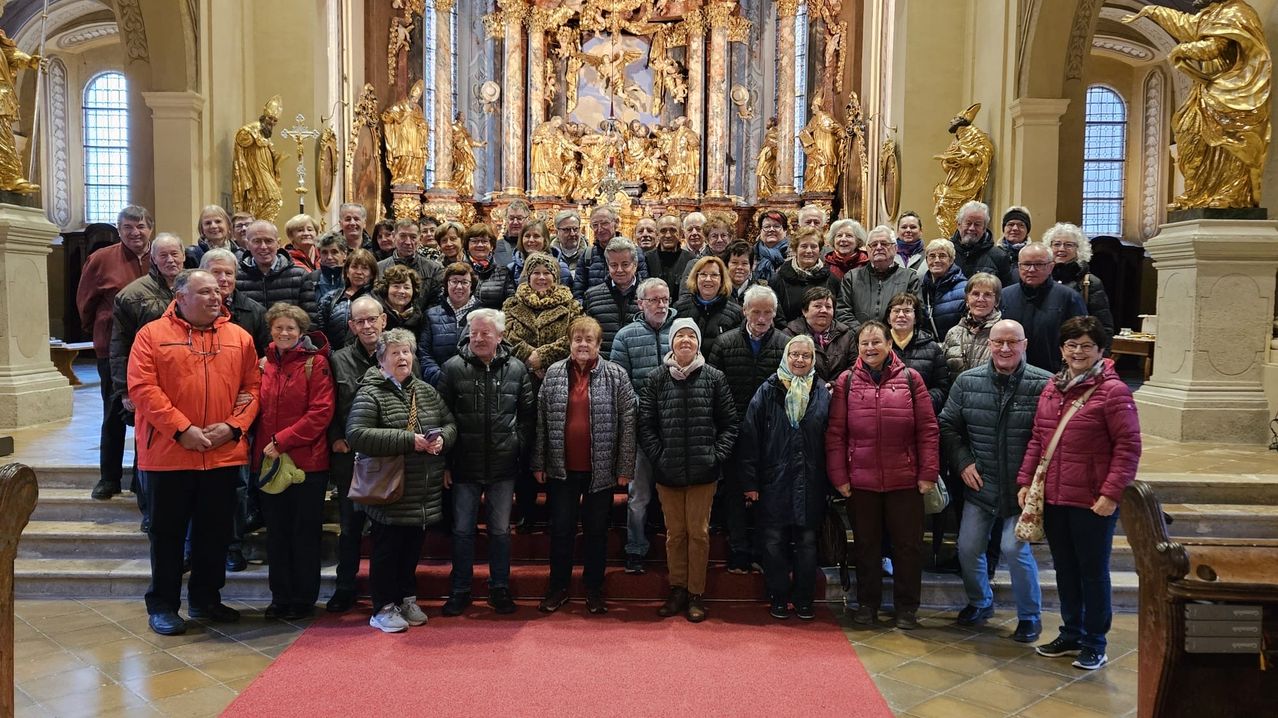 Eine Gruppe von Menschen in Winterkleidung posiert vor einem kunstvollen Kirchenaltar, mit goldenen Statuen und Buntglasfenstern im Hintergrund.