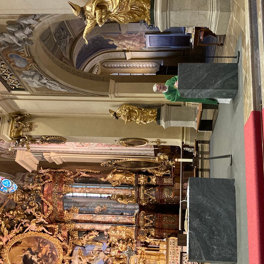 Ein reich verzierter Kircheninnenraum mit goldenen Statuen und Fresken. Ein Mann steht an einem Podium, ein roter Teppich führt zum Altar.