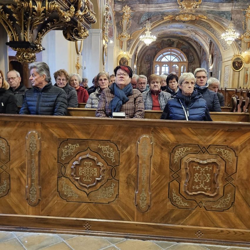 Eine Gruppe von Menschen in einer Kirche sitzt auf Kirchenbänken. Die Kirche hat kunstvoll verzierte Holzbänke, goldene Kronleuchter und Kunstwerke an den Wänden. Einige Personen tragen Brillen und Jacken.
