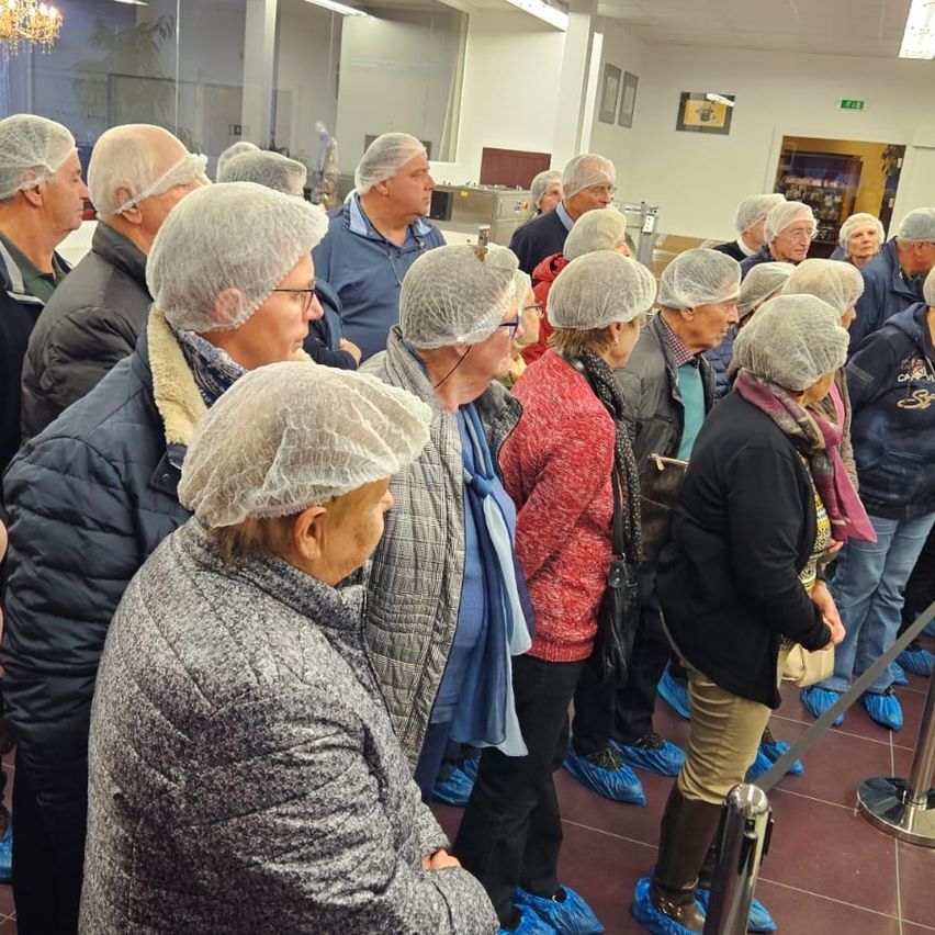 Eine Gruppe älterer Menschen mit Haarnetzen und blauen Schuhabdeckungen steht in einer Reihe in einem Raum, wahrscheinlich für eine Kochvorführung.