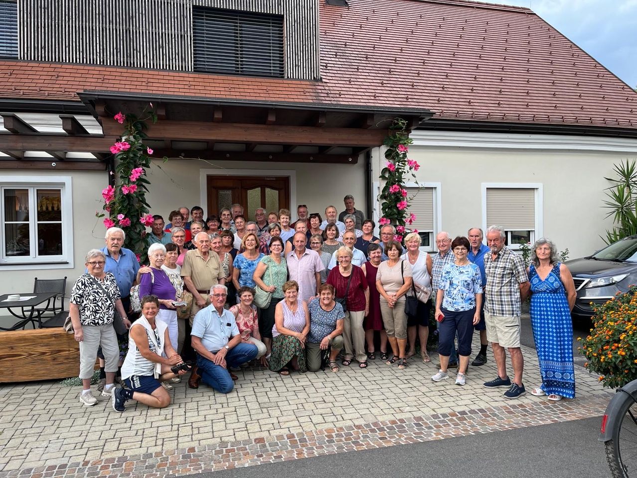 Eine Gruppe älterer Menschen posiert für ein Foto vor einem Haus mit einer Ziegelwand und einem braunen Dach.