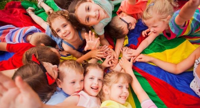 Eine Gruppe von Kindern liegt auf einer bunten Decke, lächelt und hält Händchen. Sie haben Spaß an einem sonnigen Tag.