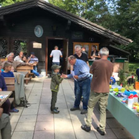Eine Gruppe von Menschen ist draußen in einem Café. Ein Junge steht zwischen zwei Männern, die sich die Hände schütteln. Andere sitzen und stehen in der Nähe.