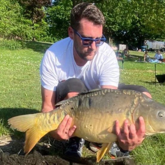 Ein Mann mit Sonnenbrille und weißem T-Shirt hält einen großen Fisch in einem grasigen Park.