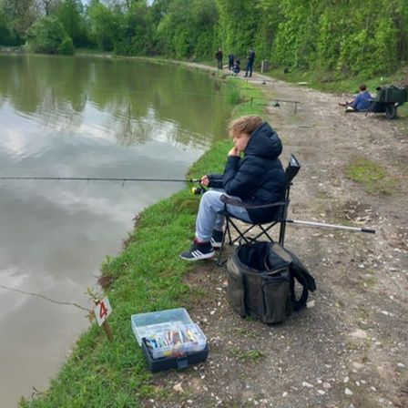 Ein junger Junge sitzt auf einem Stuhl am See und fischt mit einer Rute. In der Nähe steht ein Aufbewahrungsbehälter mit Angelködern. Im Hintergrund fischen einige Leute, und ein mit Ausrüstung beladener Schubkarren ist geparkt.