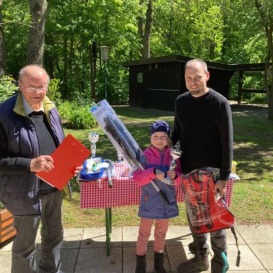 Ein Mann und ein junges Mädchen stehen nebeneinander und halten Trophäen. Hinter ihnen steht ein weiterer Mann. Sie befinden sich in einem Park mit einem Tisch vor ihnen. Auf dem Tisch befinden sich verschiedene Gegenstände. Hinter dem Tisch steht ein Gebäude.