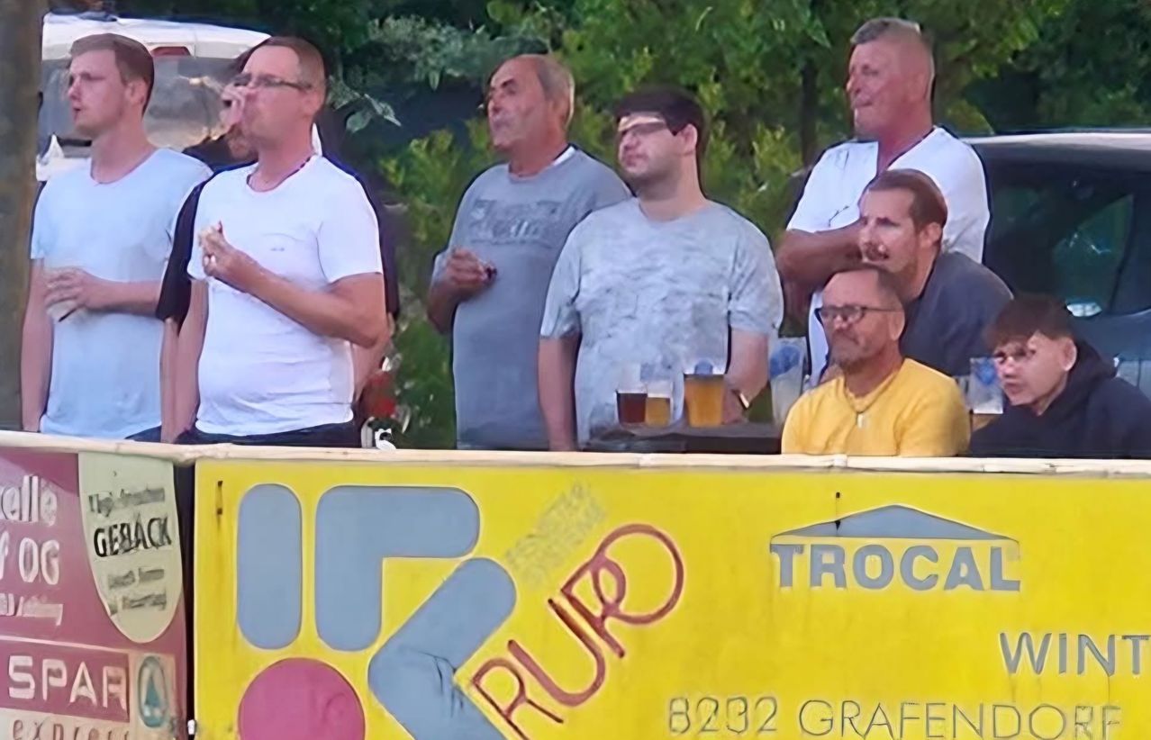 A group of men standing outdoors, some with glasses, one with a bandage on his forehead, and two with drinks on a table, with a yellow banner reading 'TROC RUPO' in the foreground.