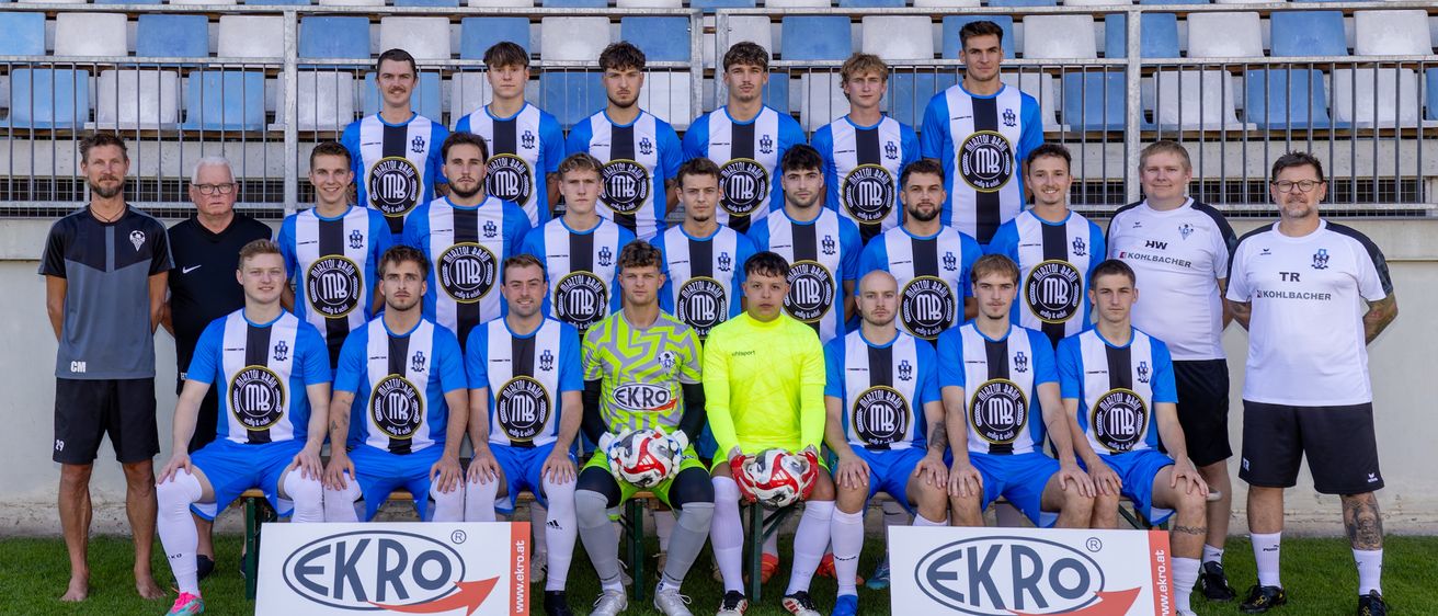 Eine Fußballmannschaft in blau-weißen Trikots mit schwarzen Streifen posiert für ein Mannschaftsfoto auf dem Spielfeld. Zwei Spieler in neongrün halten Fußballbälle. Die Mannschaft steht vor einem Banner mit der Aufschrift 'EKRO'.