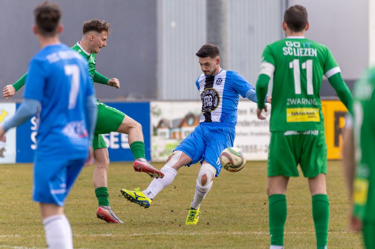 Ein Fußballspieler in blauer und weißer Uniform tritt einen Ball, während ein anderer Spieler in grüner Uniform versucht, ihn zu blockieren. Dahinter ist ein verschwommener Hintergrund eines Gebäudes und einiger Werbebanner.