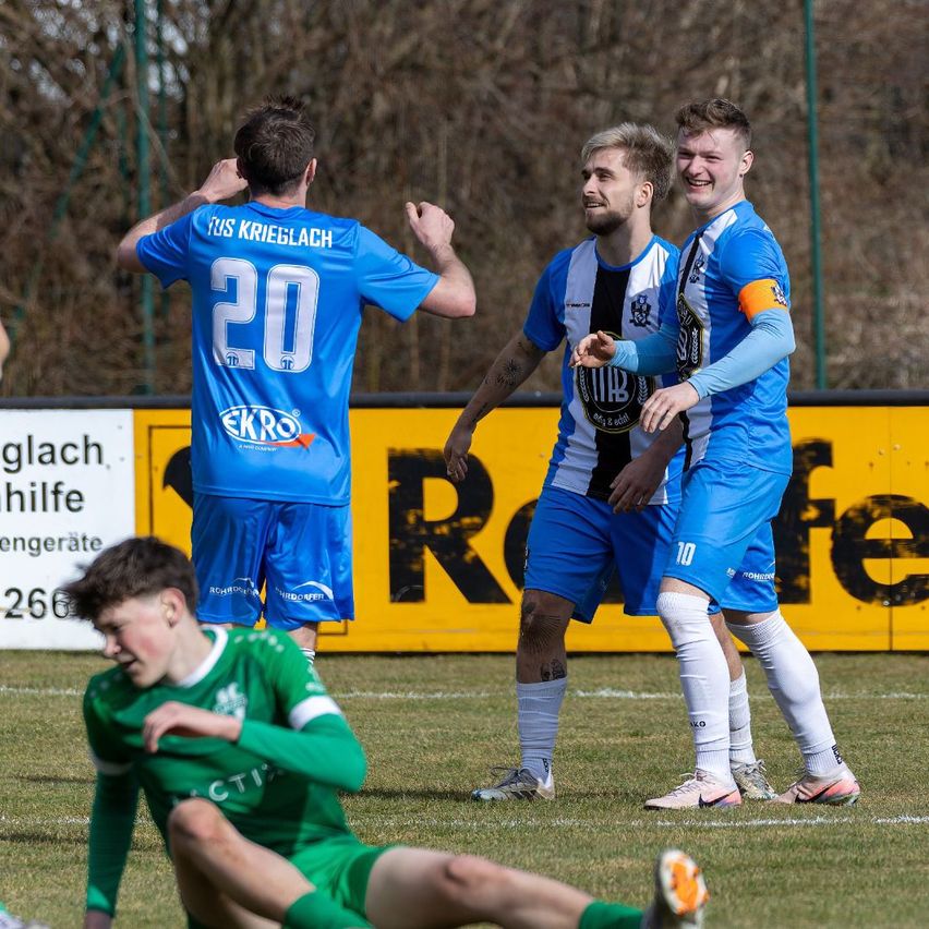 Drei Fußballspieler in blauen Trikots feiern auf einem Feld mit einem gelben Banner im Hintergrund. Ein Spieler sitzt auf dem Boden.
