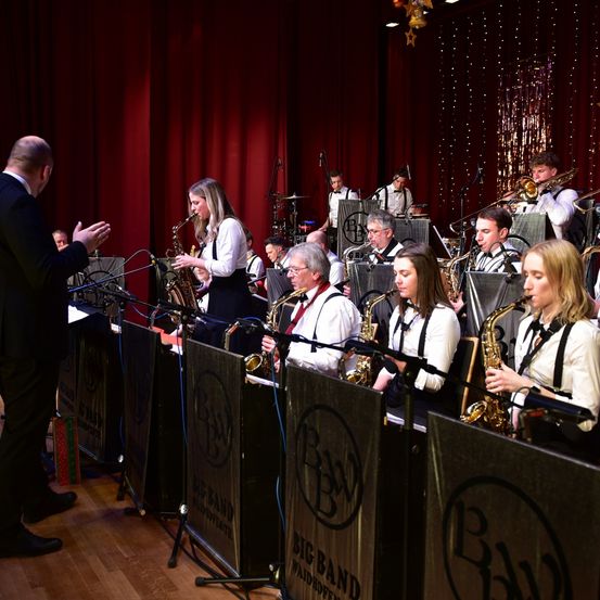 Eine Gruppe von Musikern tritt auf der Bühne auf. Eine Frau spielt Saxophon, während andere verschiedene Instrumente spielen. Das Publikum beobachtet sie von vorne. Die Bühne ist mit roten Vorhängen und Scheinwerfern dekoriert.