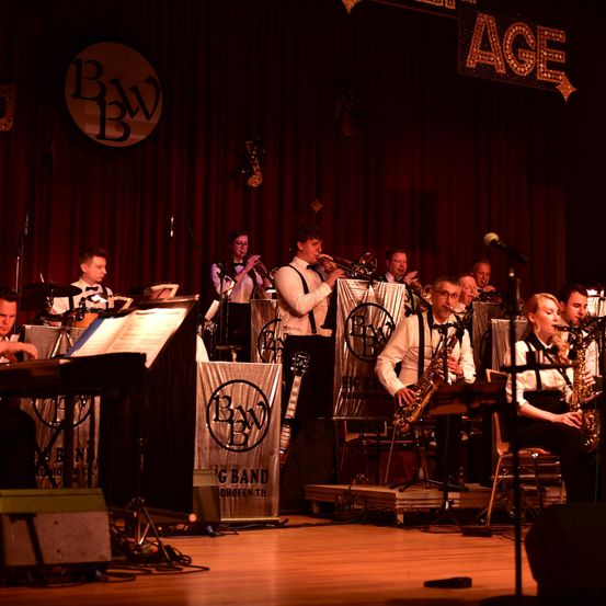Eine Jazzband spielt auf der Bühne mit Trompeten, Saxophonen und Notenblättern. Die Bühne ist rot beleuchtet, und das Publikum ist nicht sichtbar.