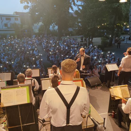 Eine Band tritt an einem Freiluftort auf, mit einem Dirigenten und Musikern auf der Bühne, die von den Taschenlampen des Publikums beleuchtet werden. Das Publikum sitzt im Halbkreis um die Bühne.