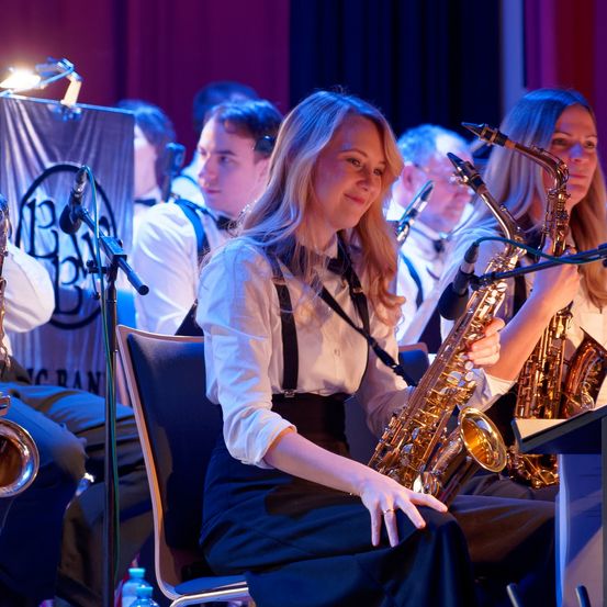 Eine Frau spielt Saxophon, während sie auf der Bühne sitzt, umgeben von einer Band. Eine weitere Frau spielt in der Nähe ein Saxophon. Die Band tritt auf der Bühne auf.