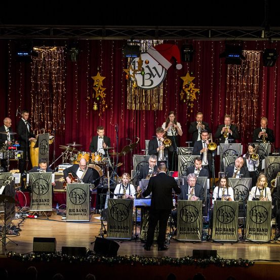 Eine Jazzband spielt auf der Bühne mit einem roten Vorhang und Weihnachtsdekorationen. Die Band besteht aus Trompeten, Posaunen, Saxophonen und einem Schlagzeug. Ein Dirigent steht vor der Band.