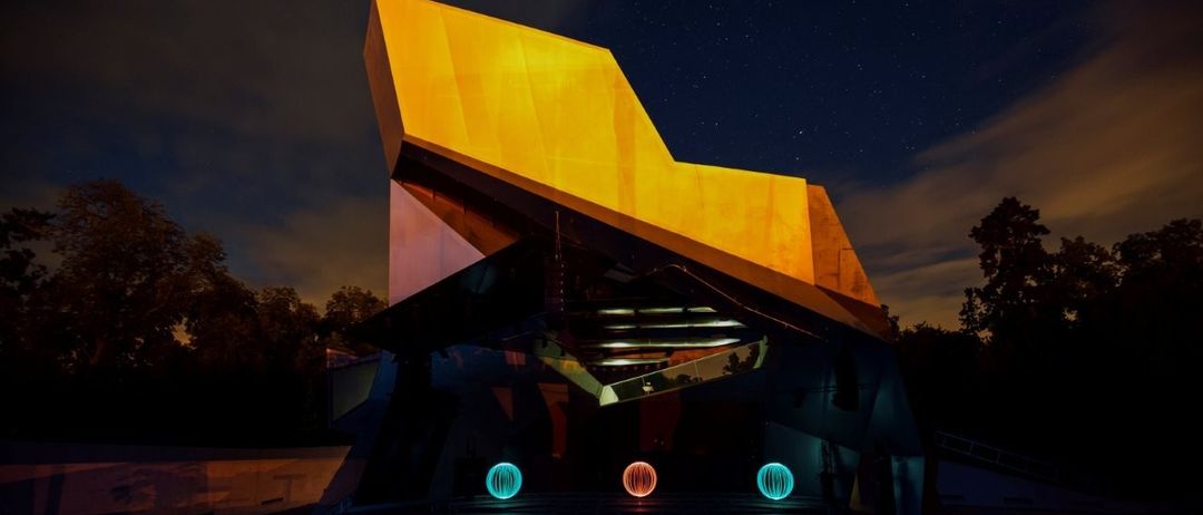 Ein modernes Gebäude mit geometrischen Formen und einem leuchtend gelben Dach ist bei Nacht beleuchtet, mit zwei leuchtend blauen Kugeln davor.