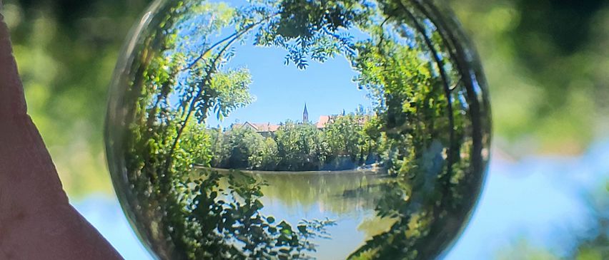 Ein Nahaufnahme eines von einer Person gehaltenen Glasballs, der eine verzerrt reflektierte Landschaft mit Bäumen, einem Fluss und einem entfernten Gebäude unter einem blauen Himmel zeigt.