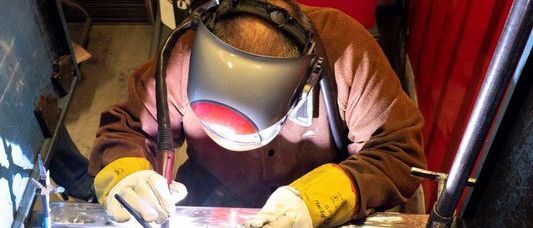 Eine Person schweißt Metall mit einer Schweißmaske und Schutzhandschuhen. Der Bereich ist mit Metallstangen und roten Lagerboxen gefüllt.