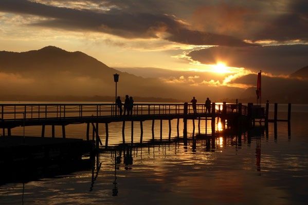 Ein silhouettierter Blick auf einen Steg mit Menschen bei Sonnenuntergang, Bergen im Hintergrund und einem bewölkten Himmel.