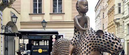 Eine Bronzestatue eines Kindes, das einen gefleckten Geparden reitet, steht vor einem Kaffeewagen mit der Marke KOFI. Im Hintergrund befinden sich Gebäude mit Fenstern und eine Straßenlampe.