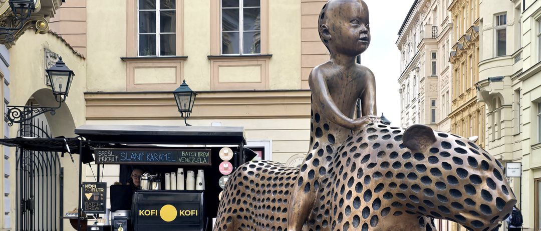 Eine Bronzestatue eines Kindes, das einen gefleckten Geparden reitet, steht vor einem Kaffeewagen mit der Marke KOFI. Im Hintergrund befinden sich Gebäude mit Fenstern und eine Straßenlampe.