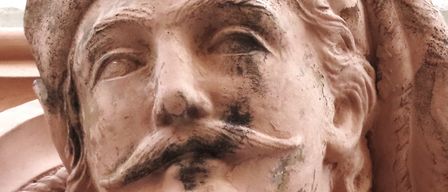 Eine Nahaufnahme einer verwitterten Statue, die einen bärtigen Mann mit Schnurrbart zeigt, der einen Hut trägt und auf seiner Oberfläche feine Details aufweist.
